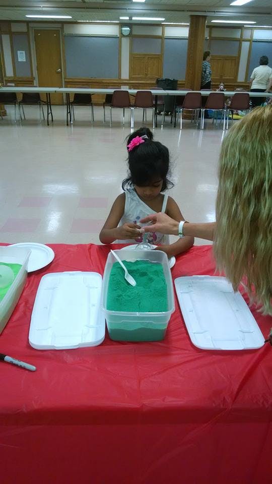 Child at a table with green sand, interacting with an adult's hand