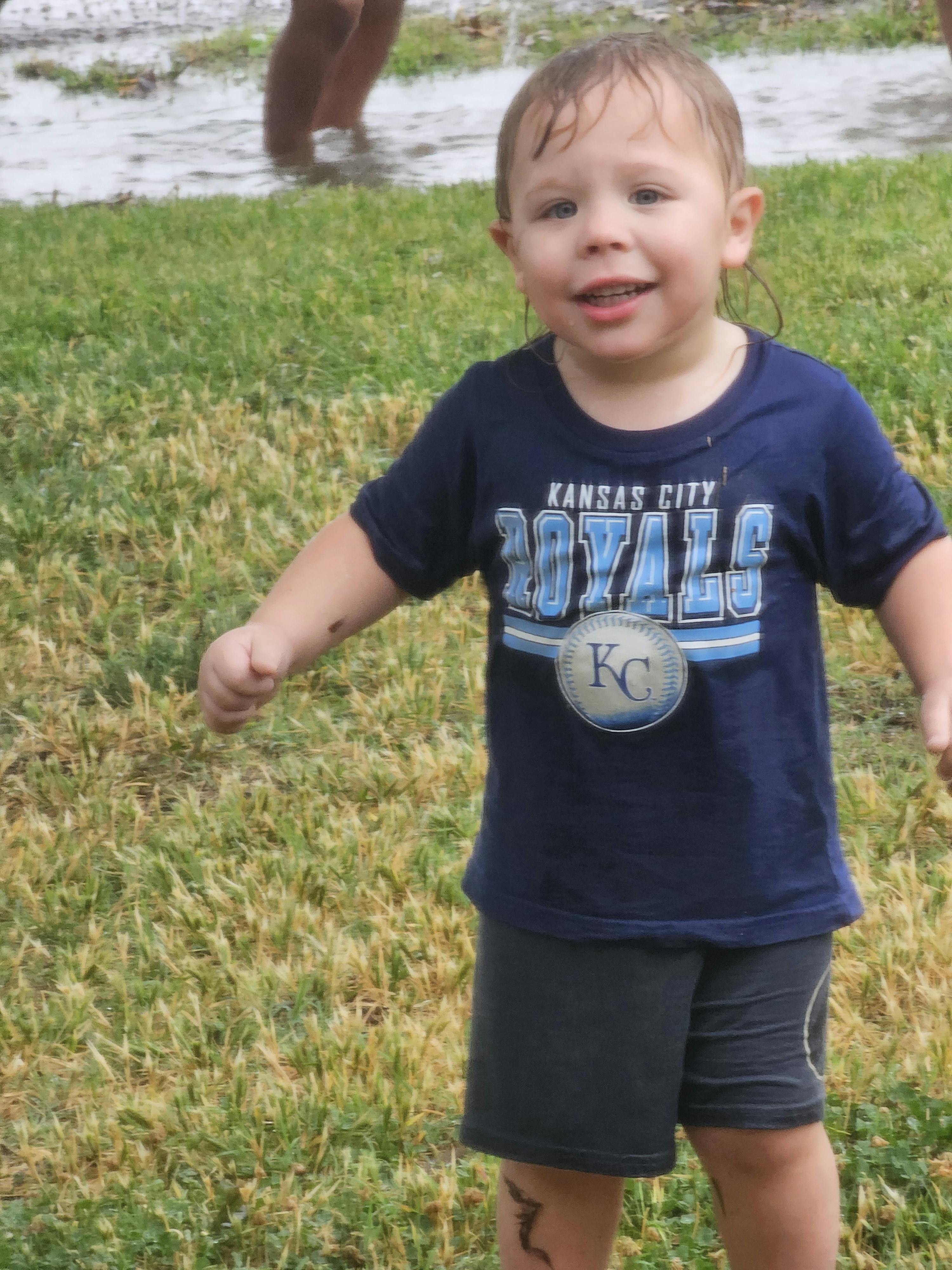 Child in a Kansas City Royals shirt standing in a grassy area with water
