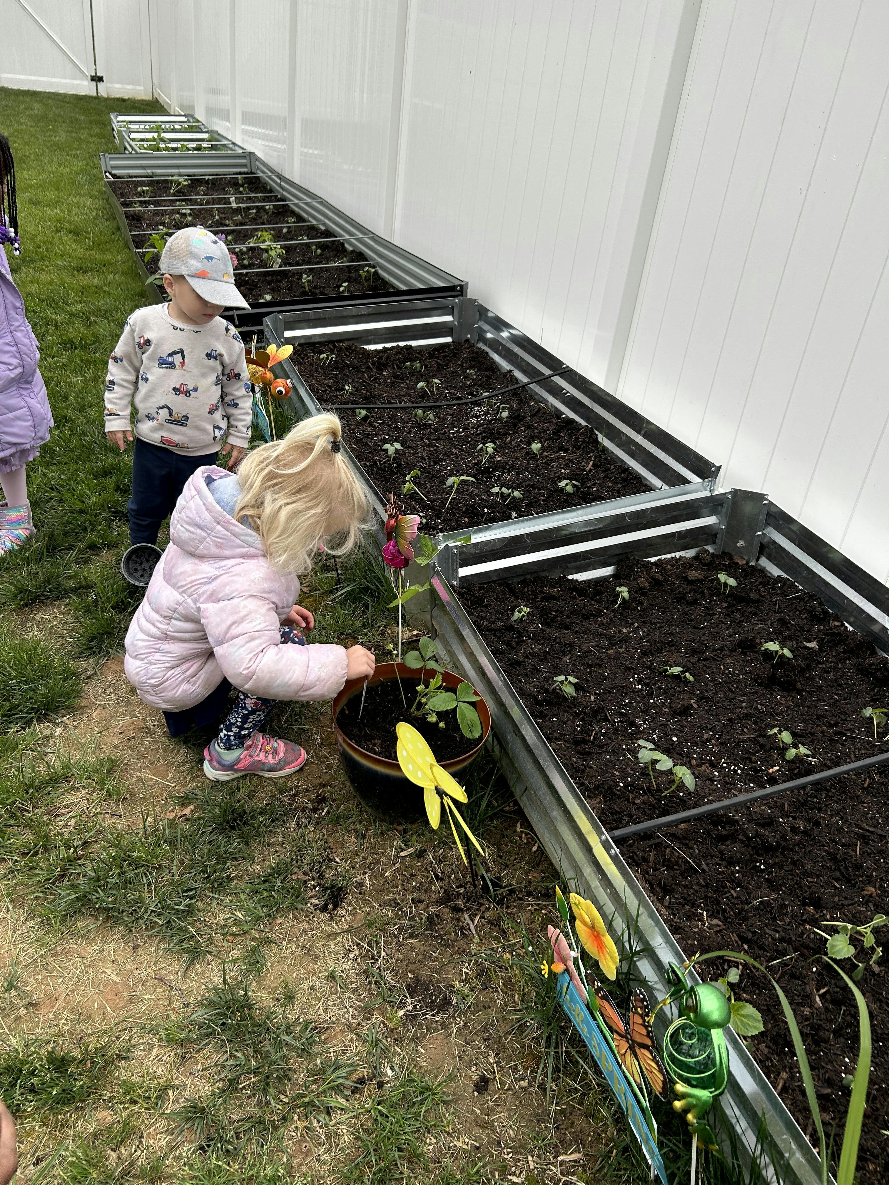 Child crouching next to a plant pot, another child standing nearby, beside garden beds
