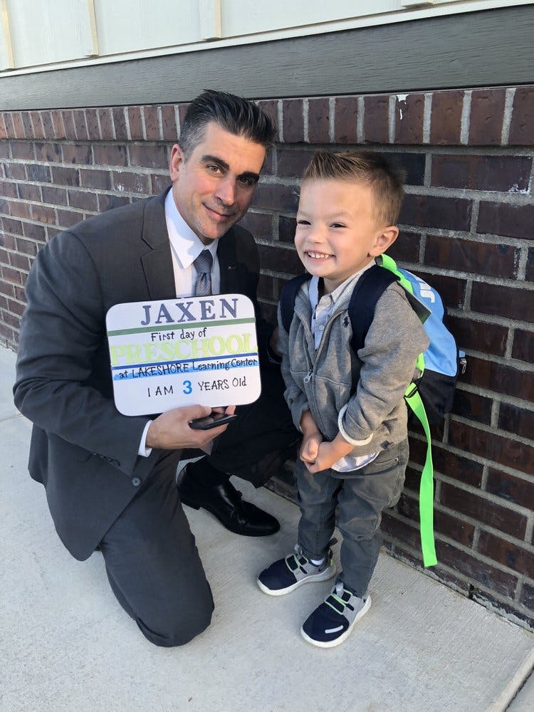 Person in suit kneeling next to a child with a backpack, holding a sign that says "JAXEN First day of SCHOOL I AM 3 YEARS OLD"