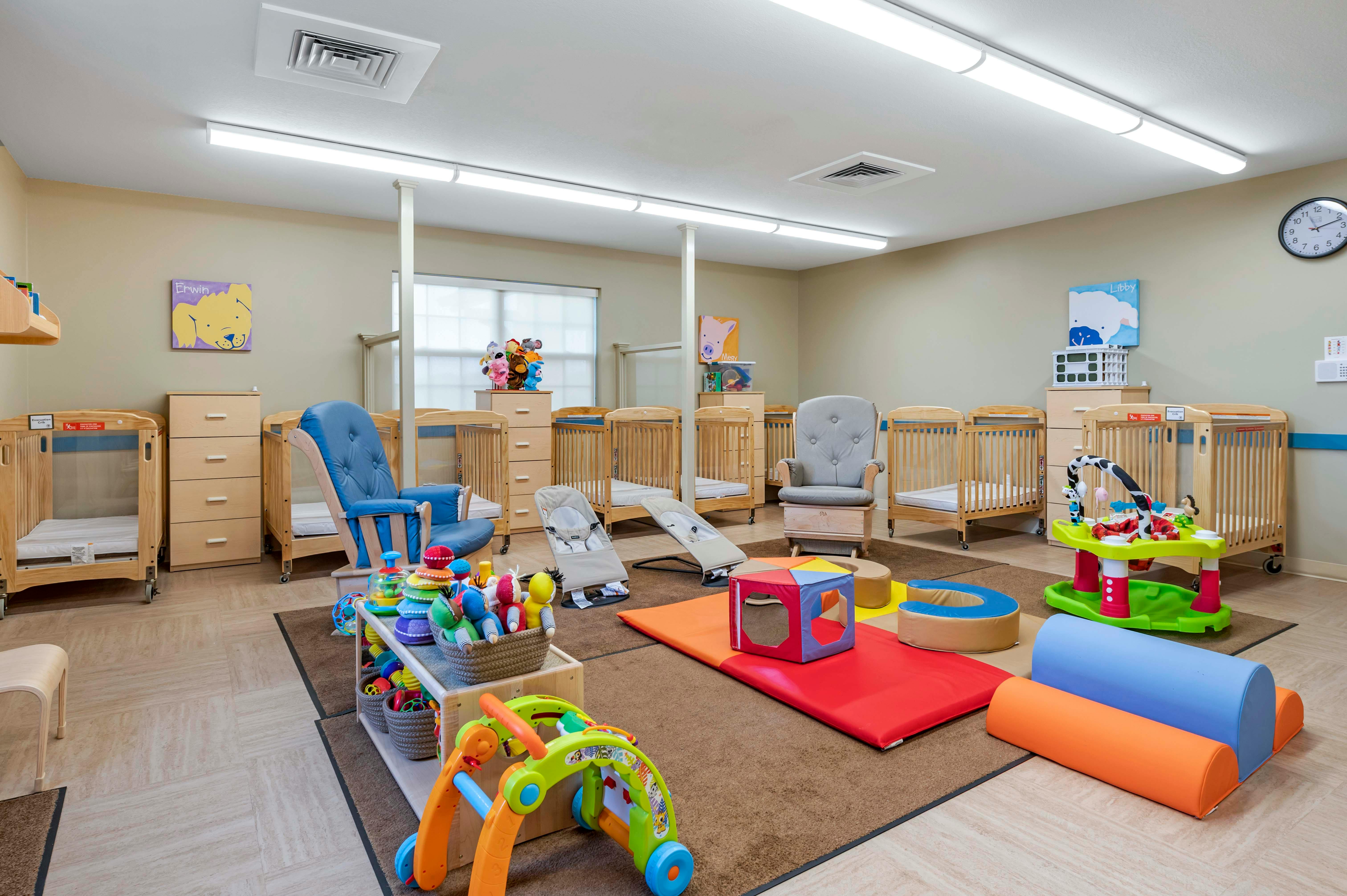 cribs in a daycare room with chairs, play mats, and baby toys