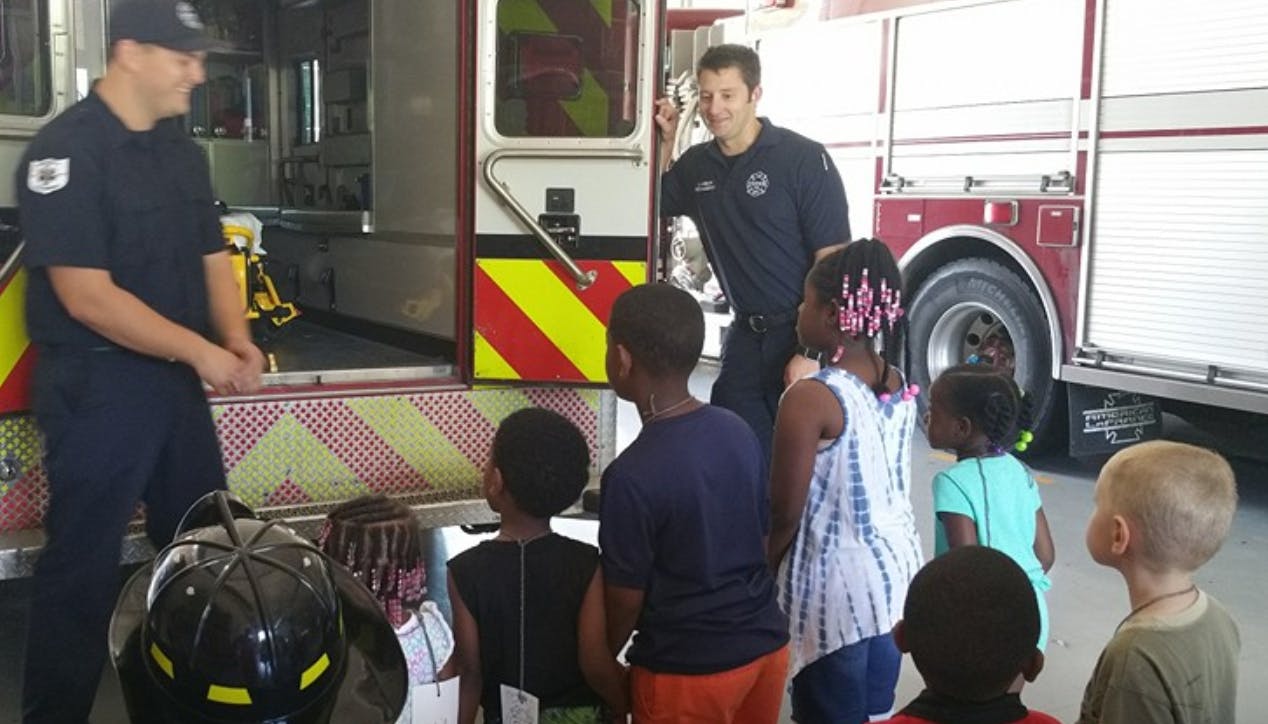 Two people in uniform with children near a fire truck