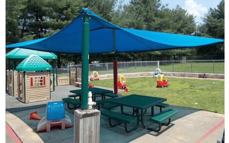 Playground with playhouses, picnic tables, and toy cars under a large blue shade