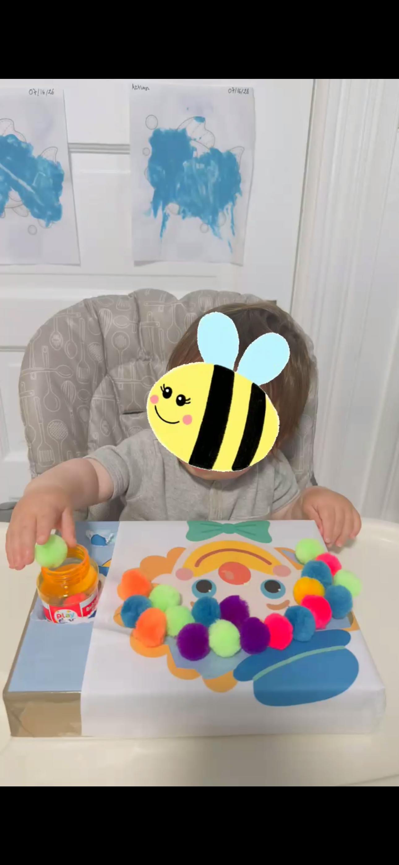 Child in a high chair playing with a clown craft and colorful pom poms