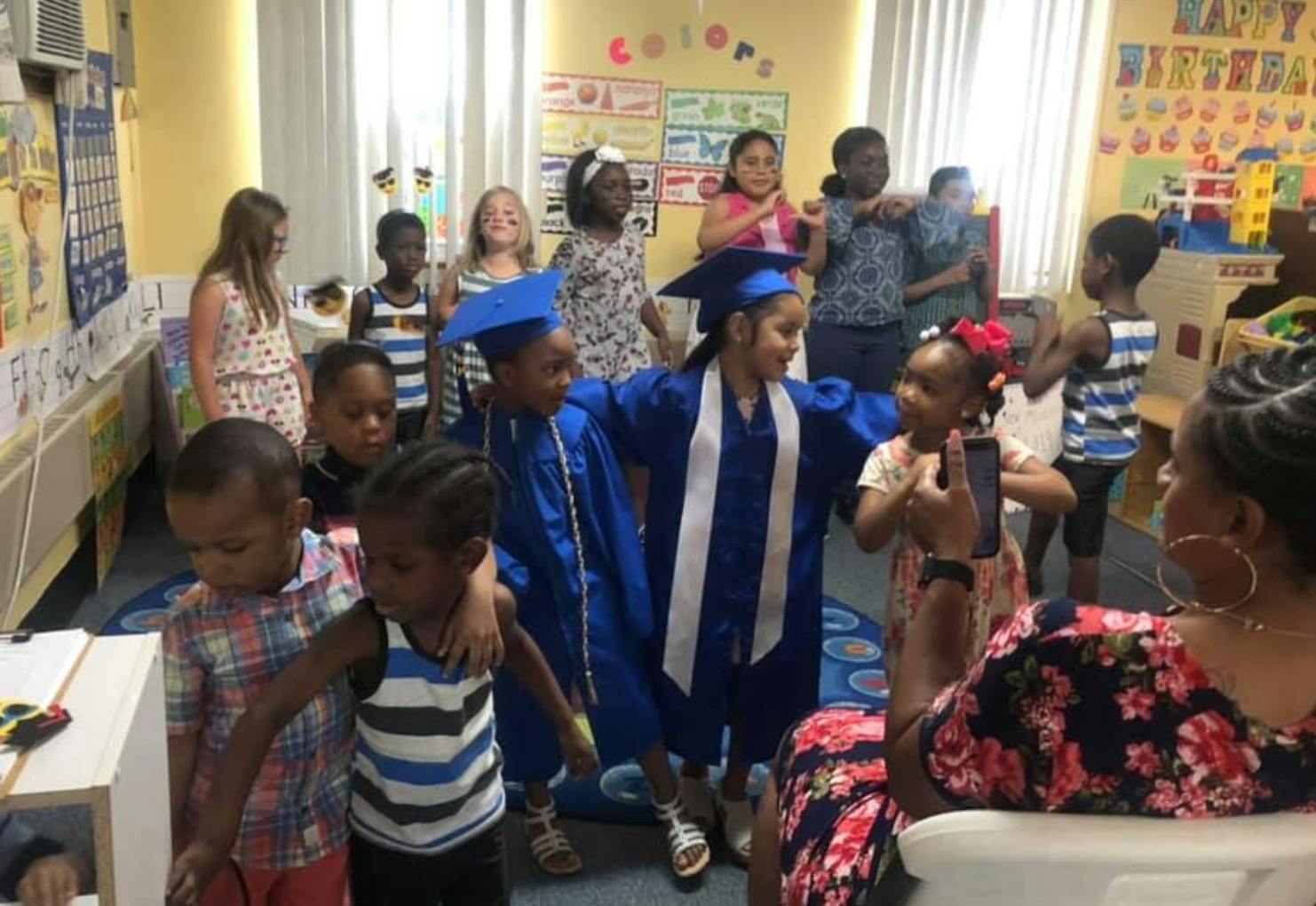 Children in graduation caps and gowns with other children and adults in a classroom