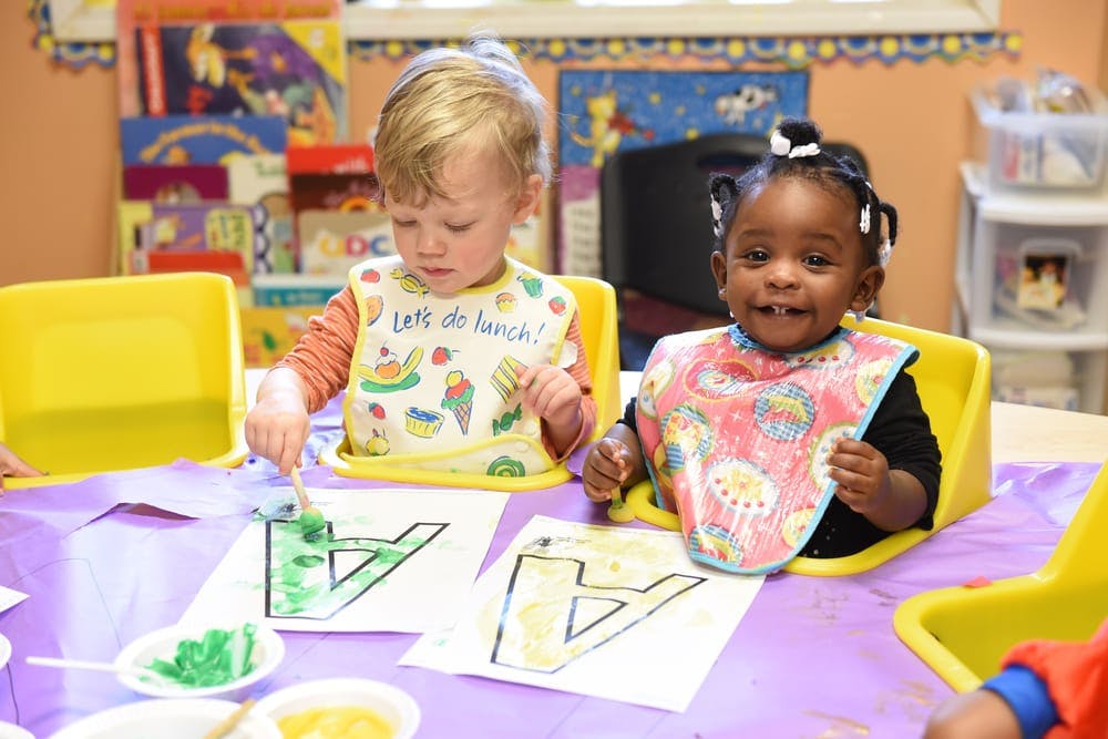 Two children in bibs painting letter A outlines at a table