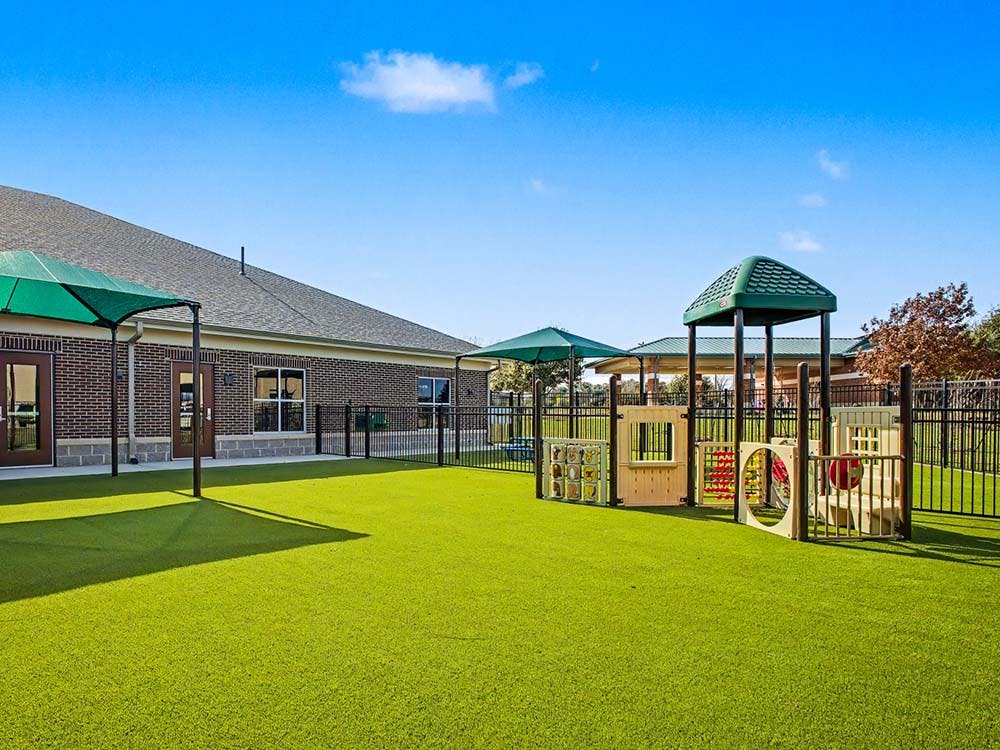 Playground with green turf, umbrellas, and play structures outside a brick building