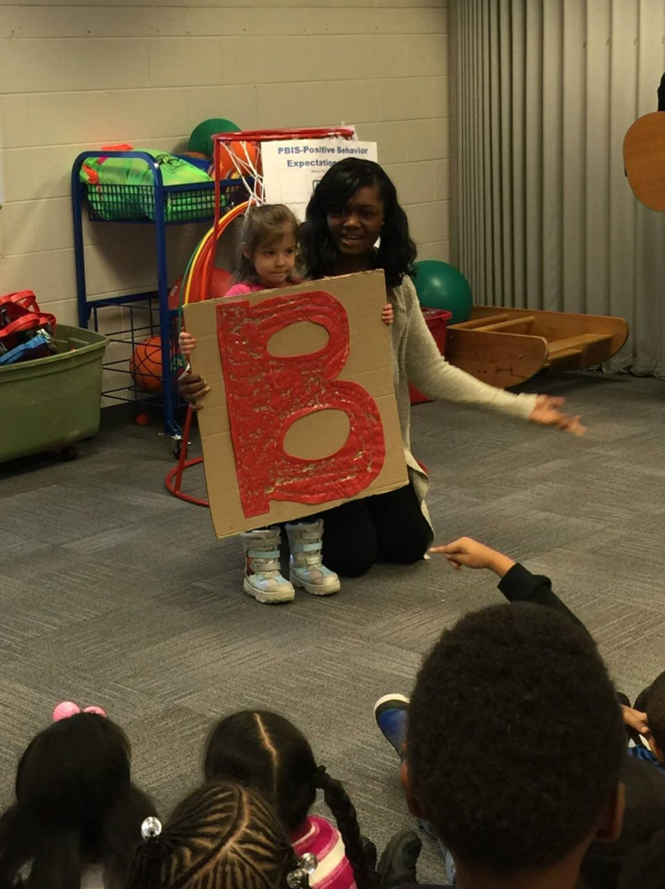 Person and child holding a large letter B in front of seated children