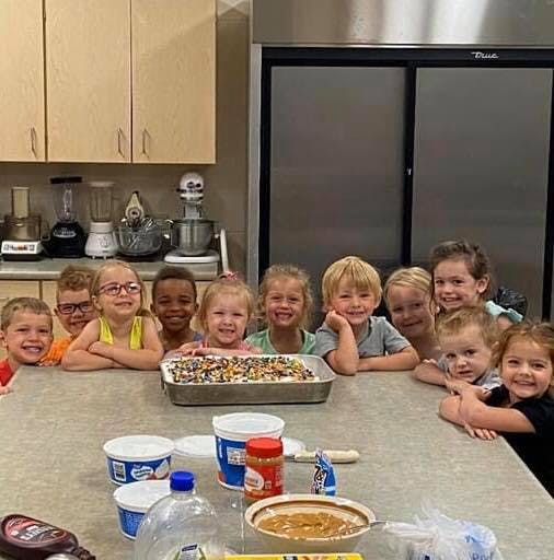 Group of children gathered around a table with a dessert in a kitchen