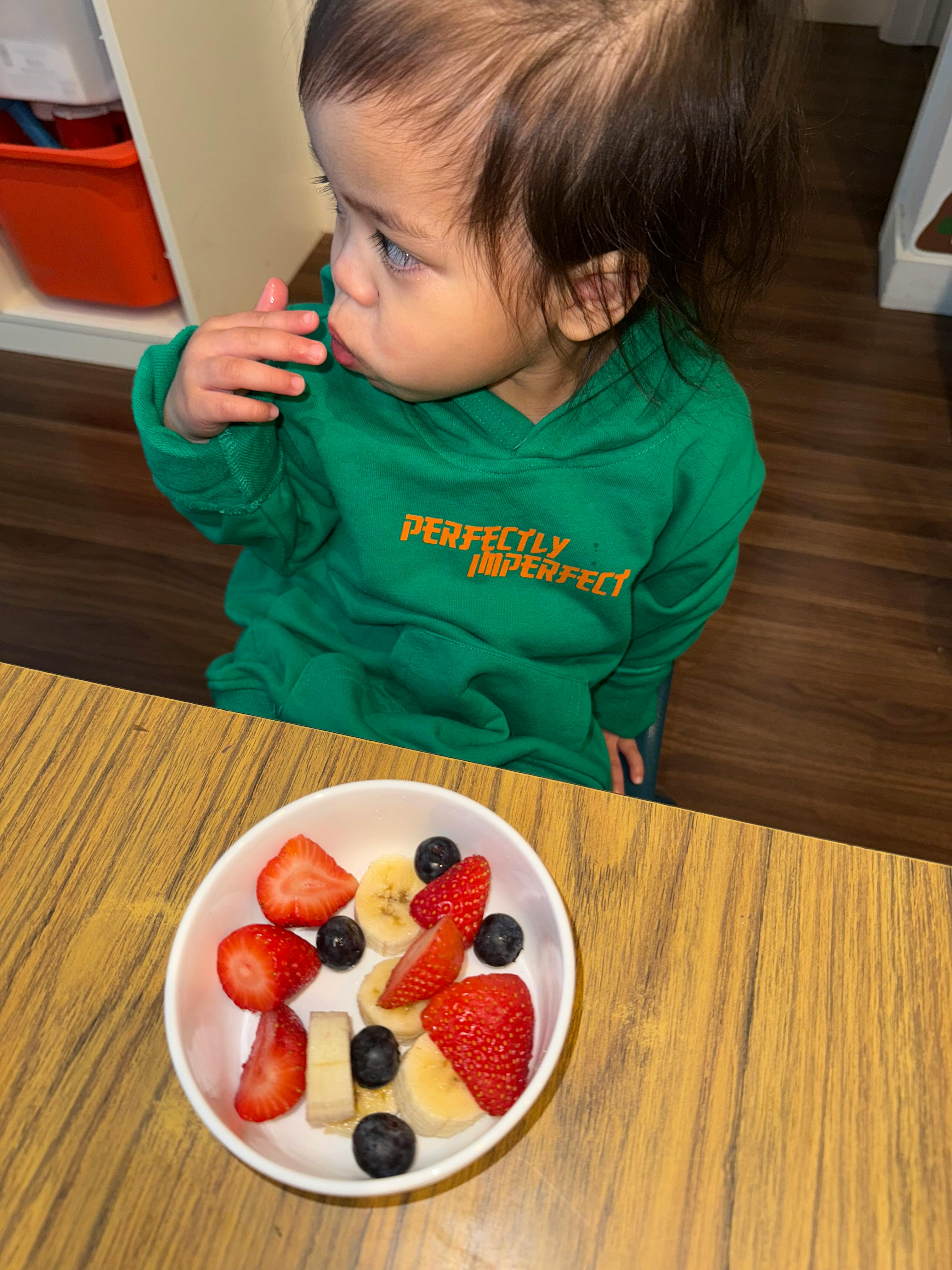 Child in a green hoodie looking at a bowl of fruit
