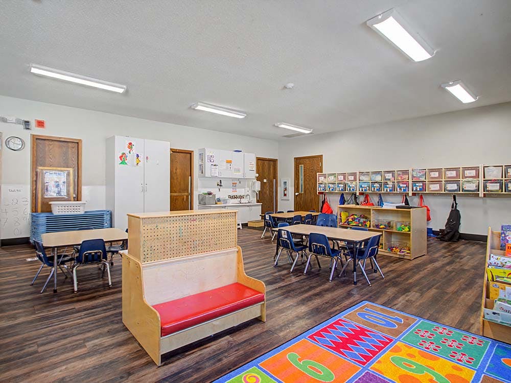 Classroom with small tables, chairs, colorful rug, and storage shelves