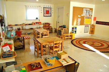 Classroom with small wooden tables and chairs, shelves with learning materials