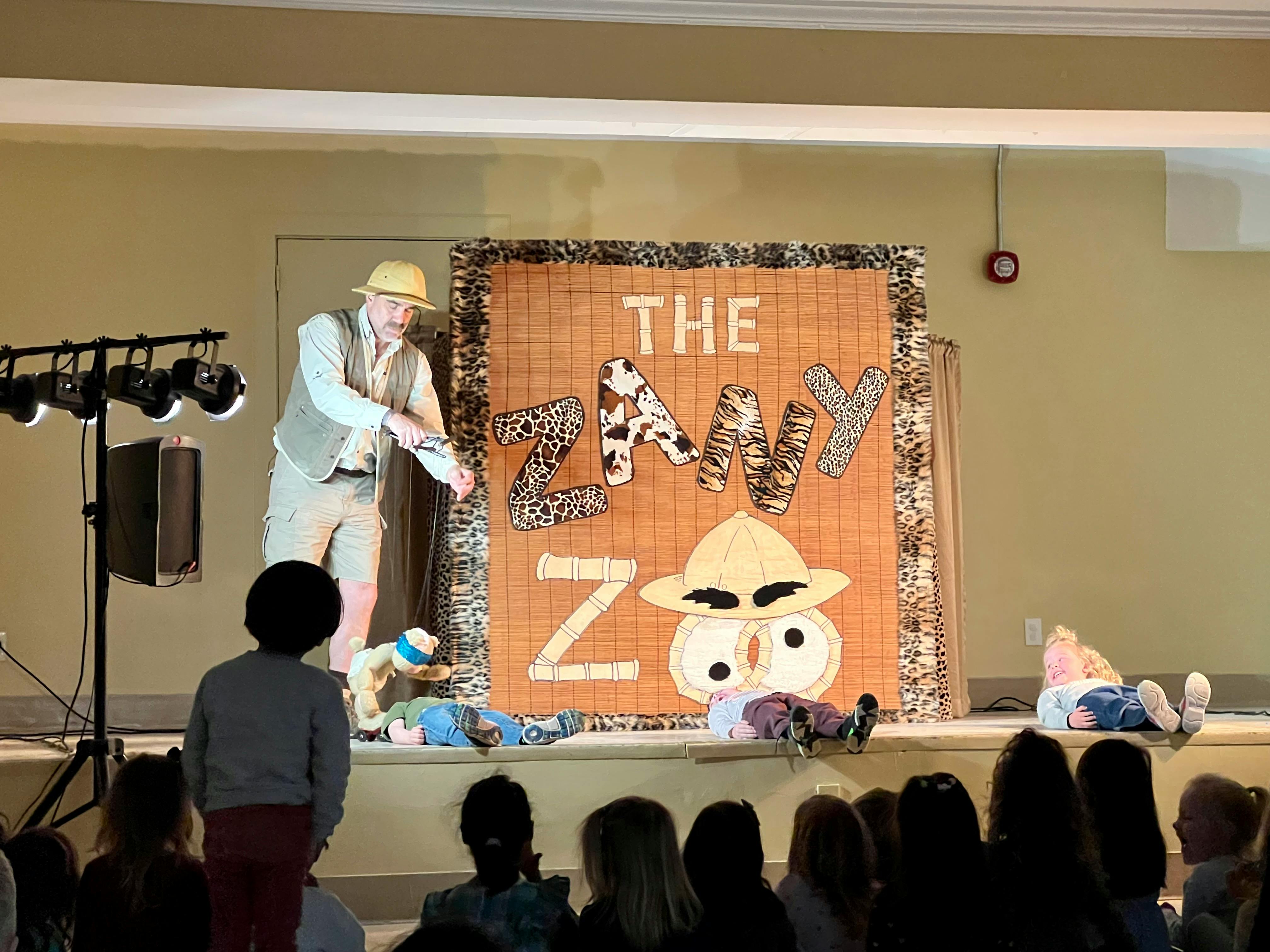 Person on stage performing in front of an audience with a "The Zany Zoo" backdrop