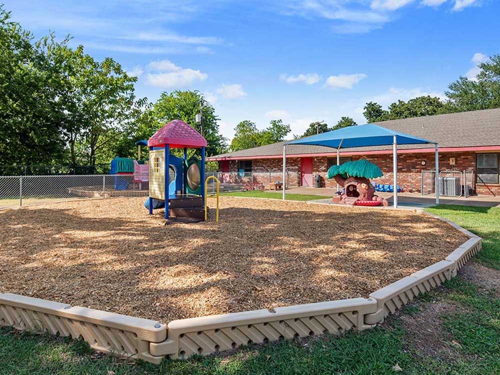 Outdoor playground with wood chips, colorful play structures, and a brick building
