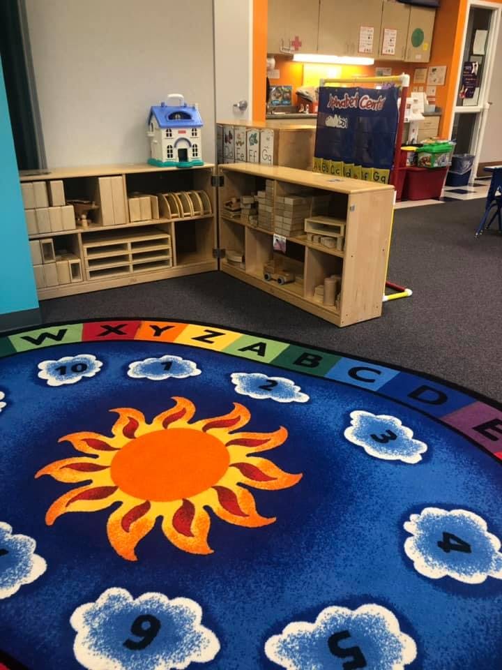 Classroom with a sun and clouds rug, wooden shelves with blocks and toys