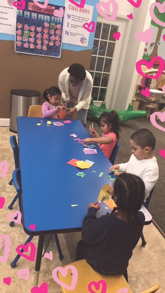 Adult helping children with paper crafts at a blue table in a classroom