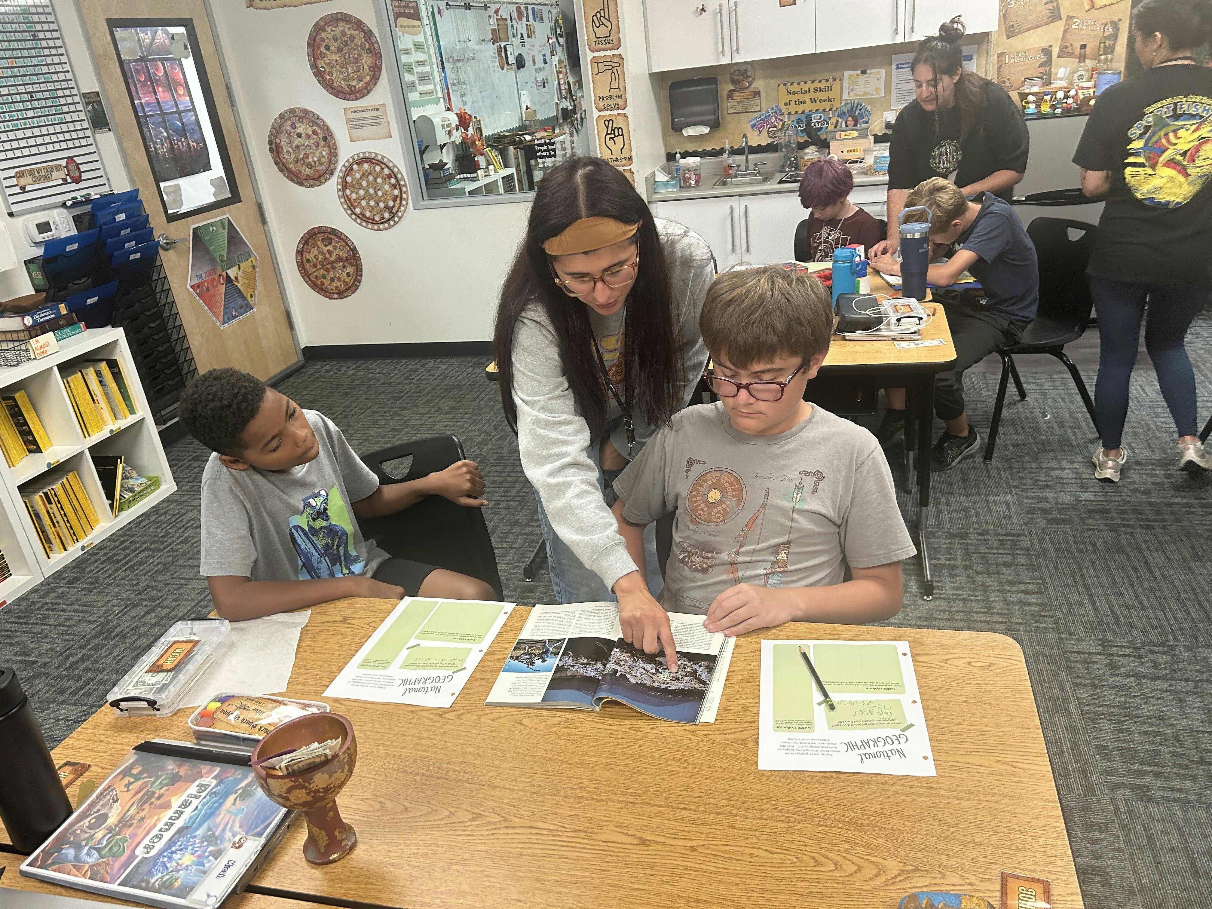 A person helping two students with a magazine in a classroom