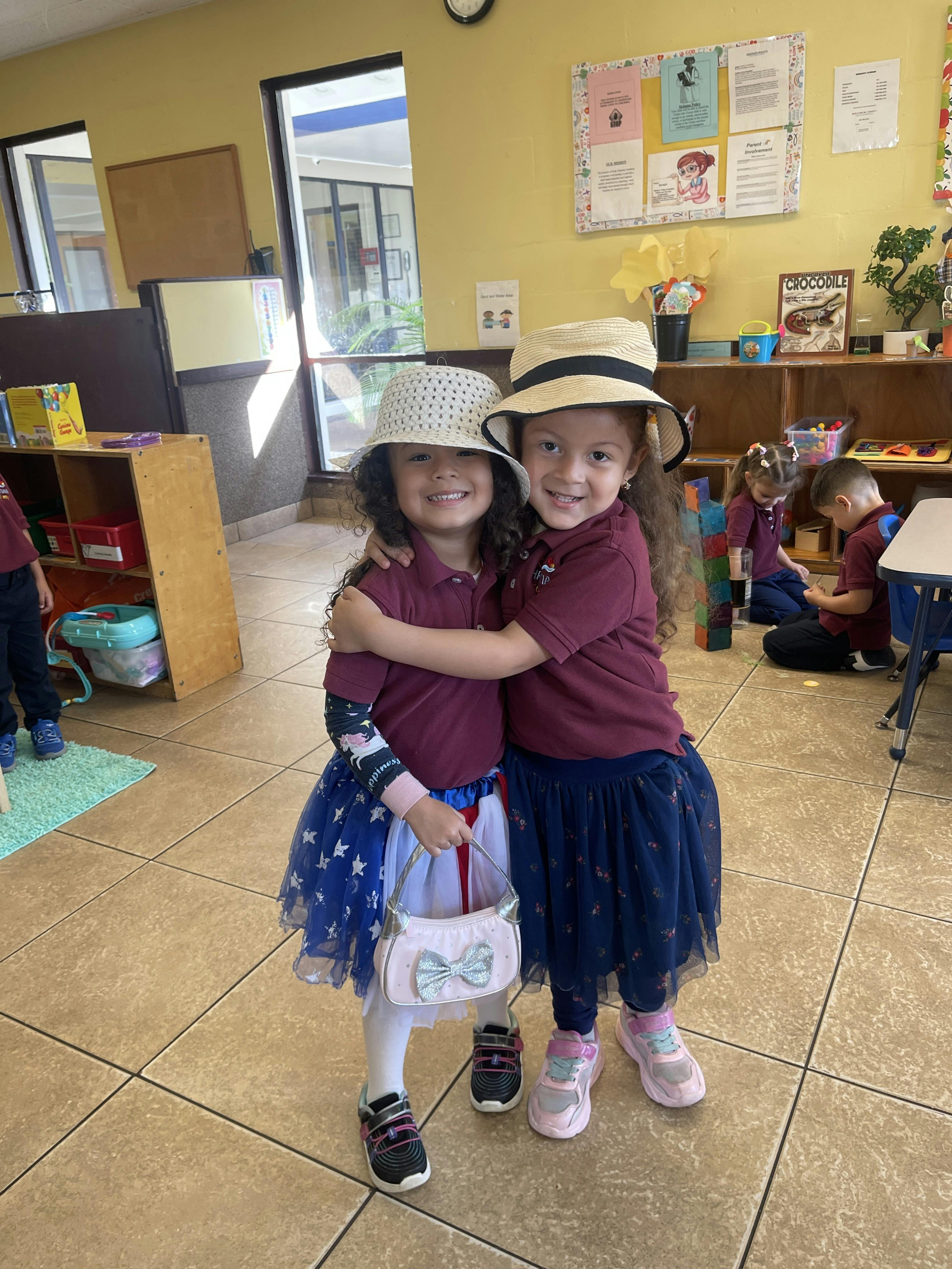 Two children wearing hats and matching outfits hugging in a classroom