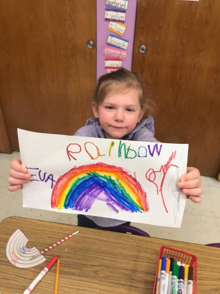 Child holding up a drawing of a rainbow with the word "rainbow" written above it