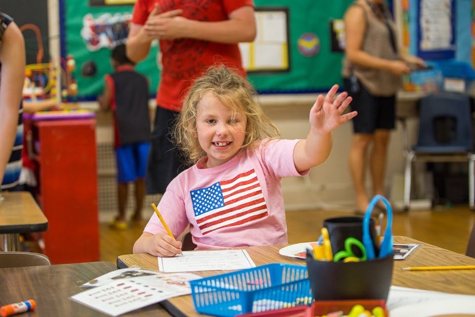 Child in a classroom raising a hand while writing