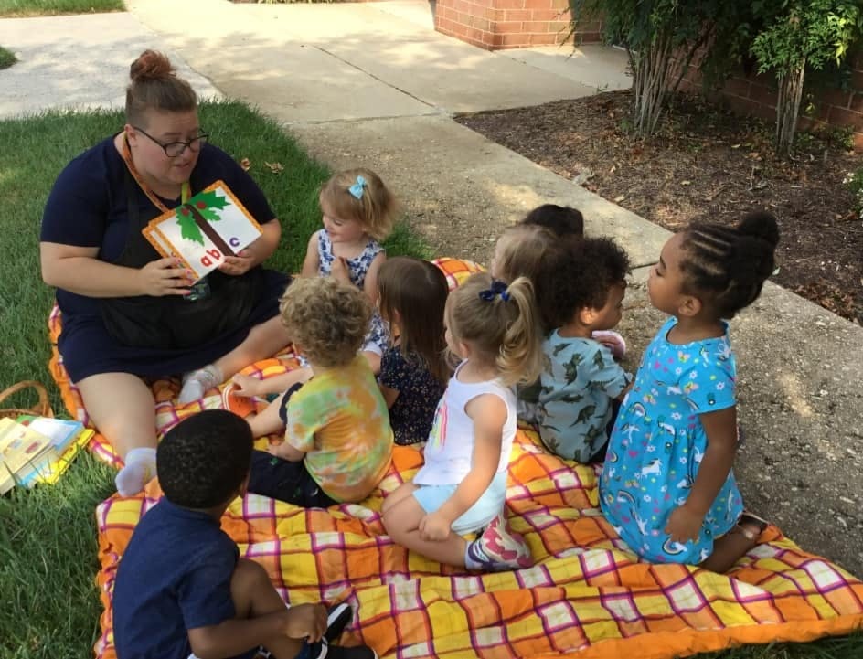 Person reading a book to a group of children sitting on a blanket outdoors