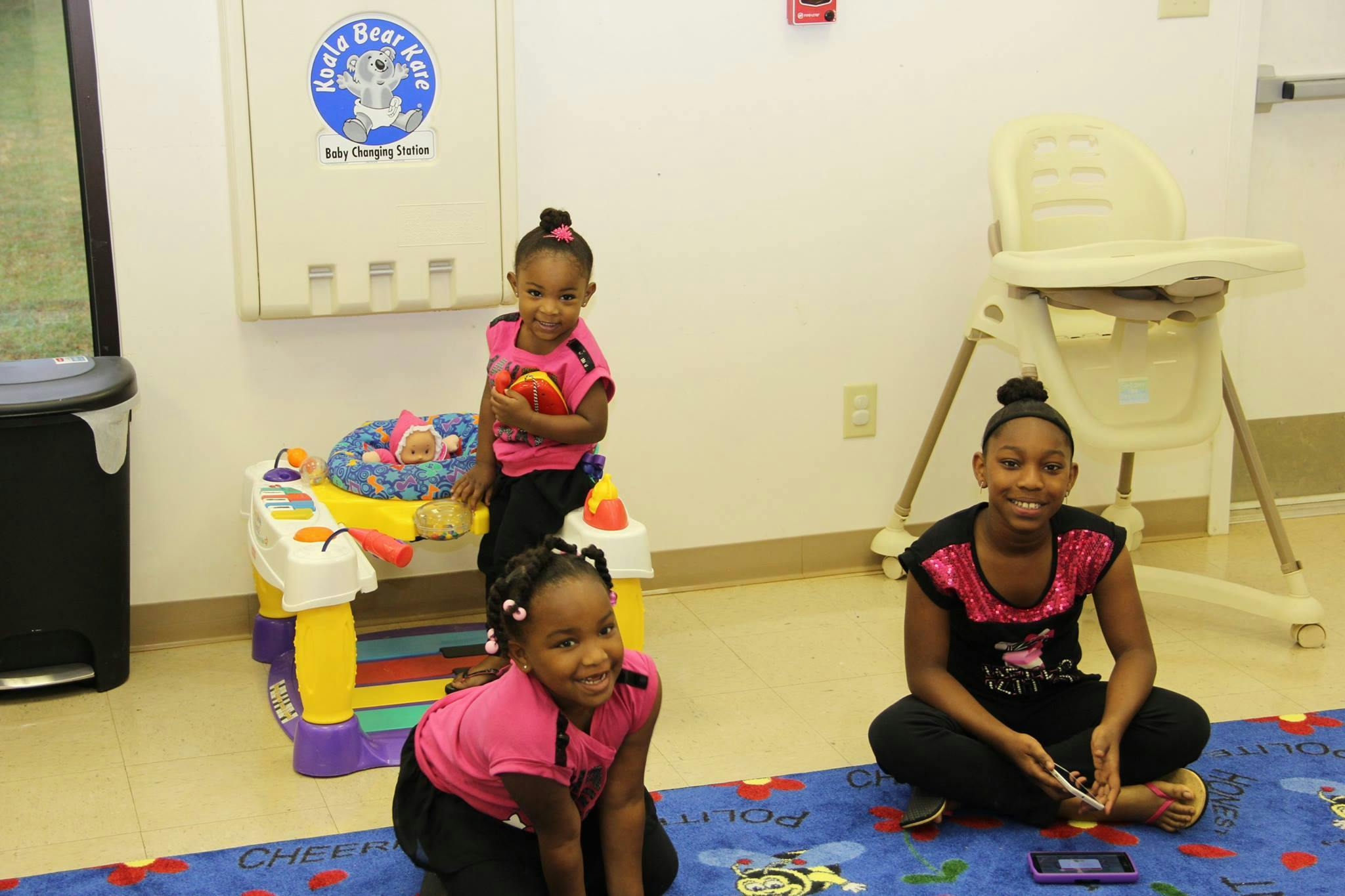 Three children in a room, one standing, one kneeling, one sitting on a rug