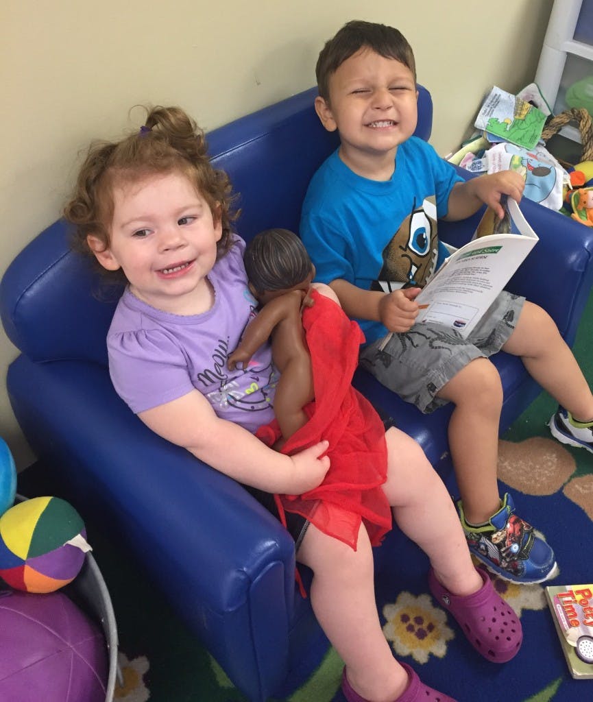 Two children sitting on blue chairs, one holding a doll and the other reading a book