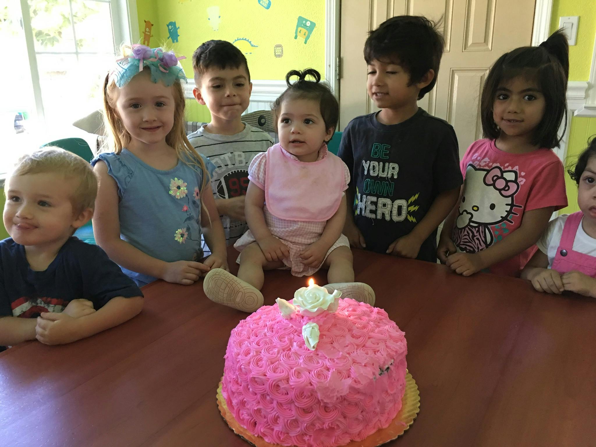 Children gathered around a pink birthday cake with a lit candle