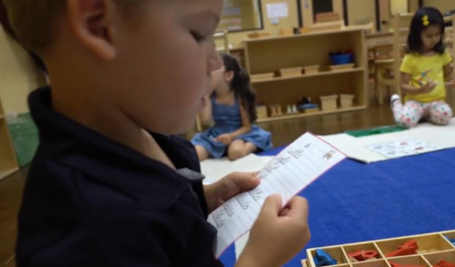 Child holding a paper in a classroom with other children in a classroom