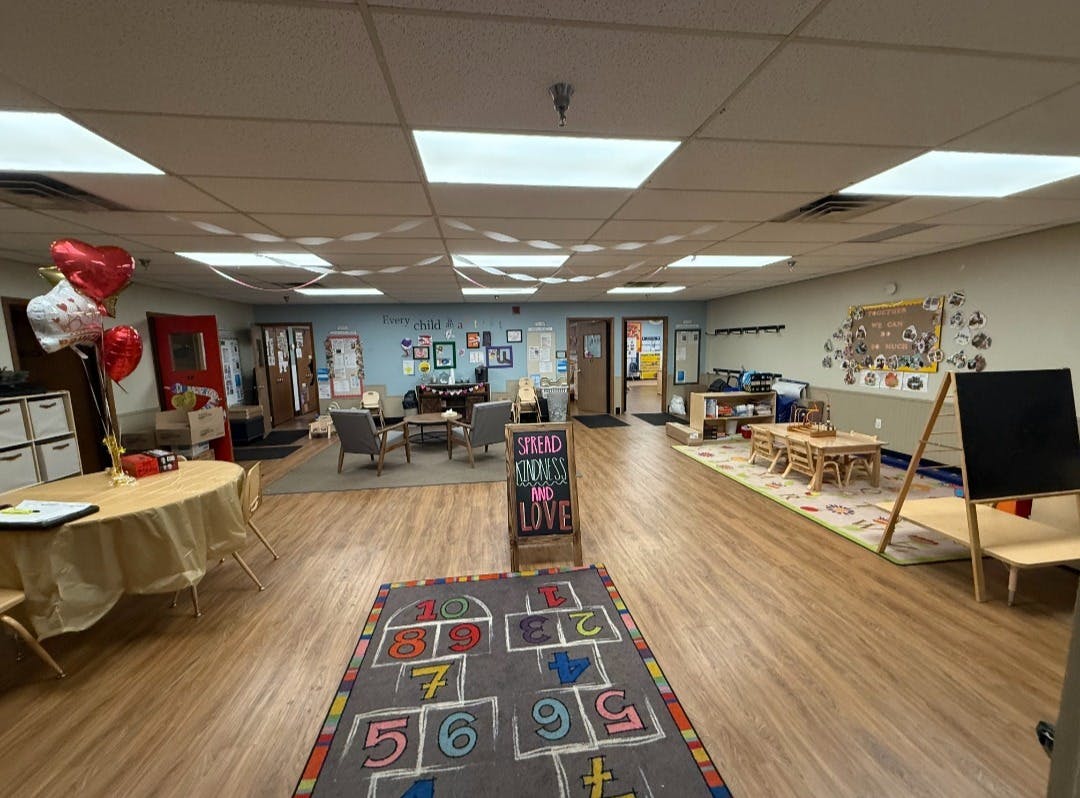 An empty childcare center or classroom with small tables, chairs, and a hopscotch rug