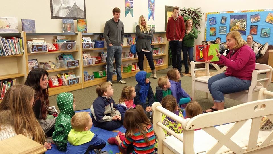 Person reading a book to a group of children and adults in a room with bookshelves