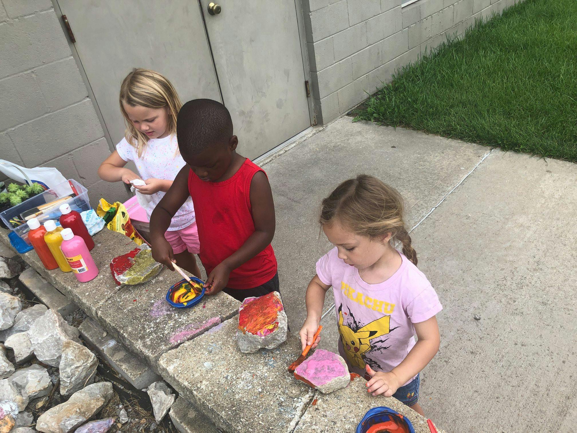 Three children painting rocks outdoors