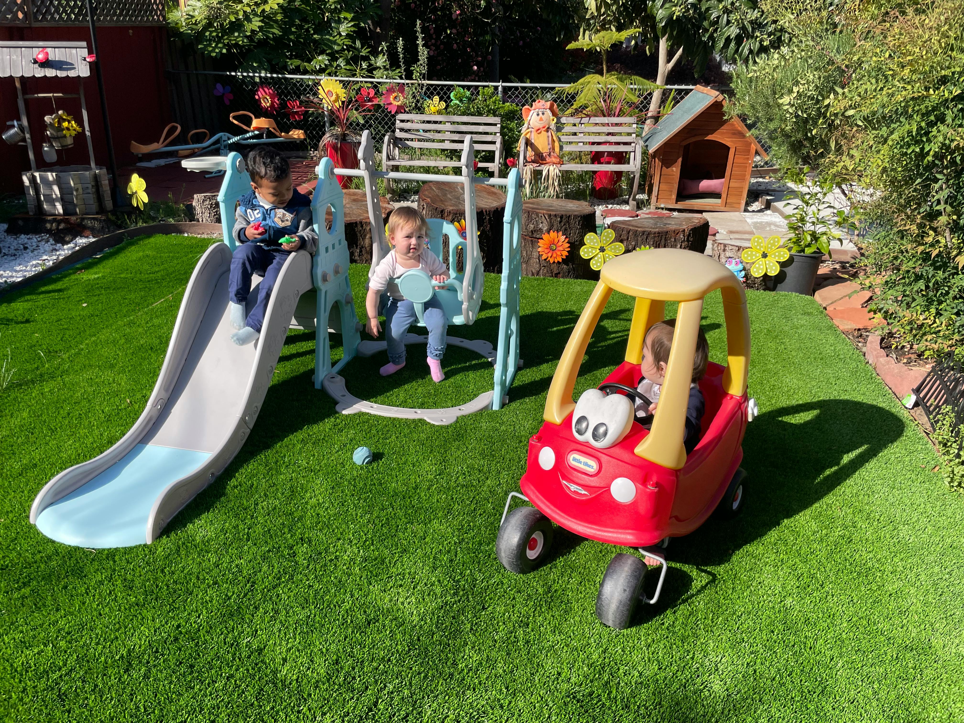 Children playing on a slide, swing, and toy car in a backyard