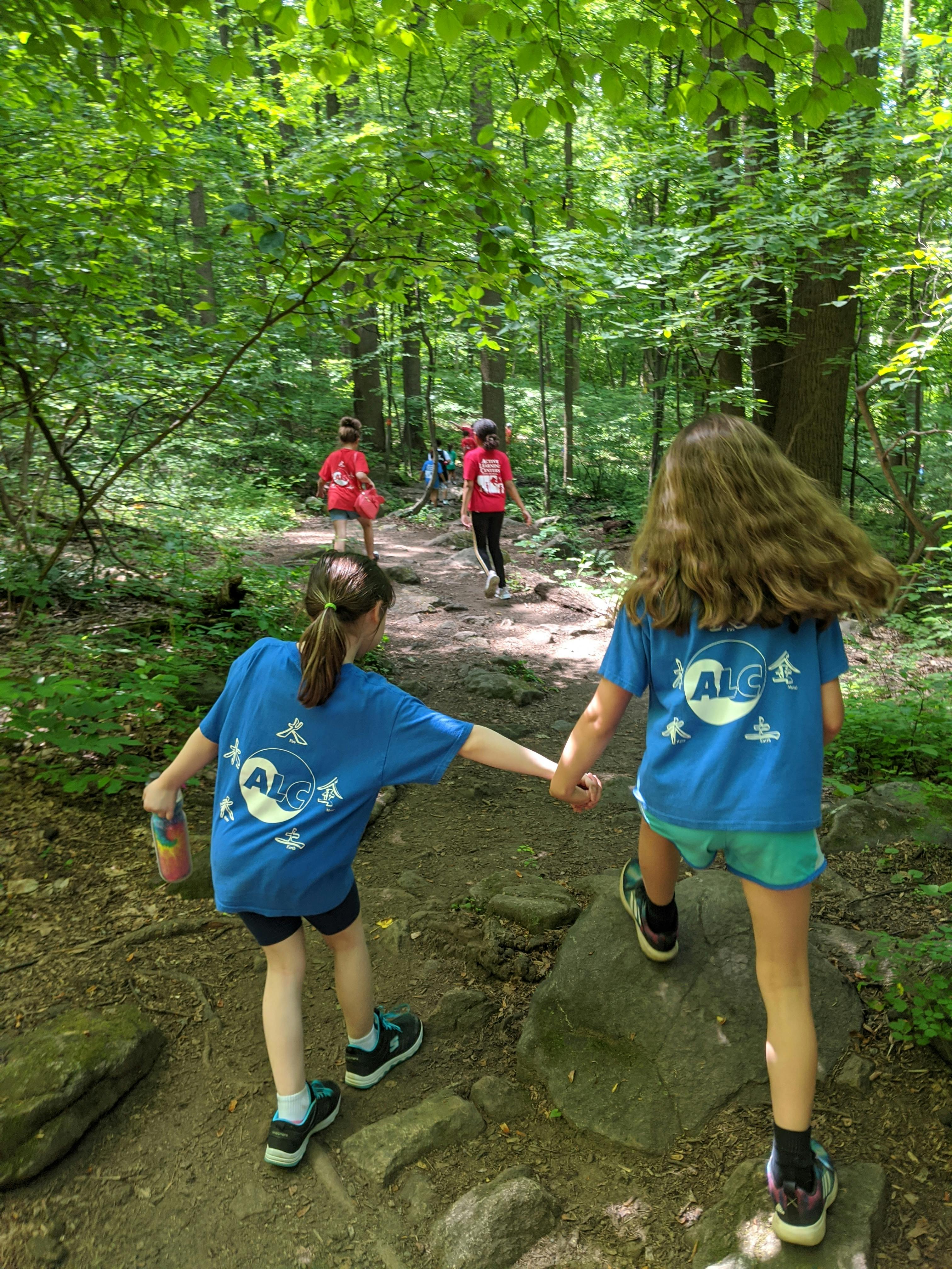 Two children holding hands on a wooded path, other children in background
