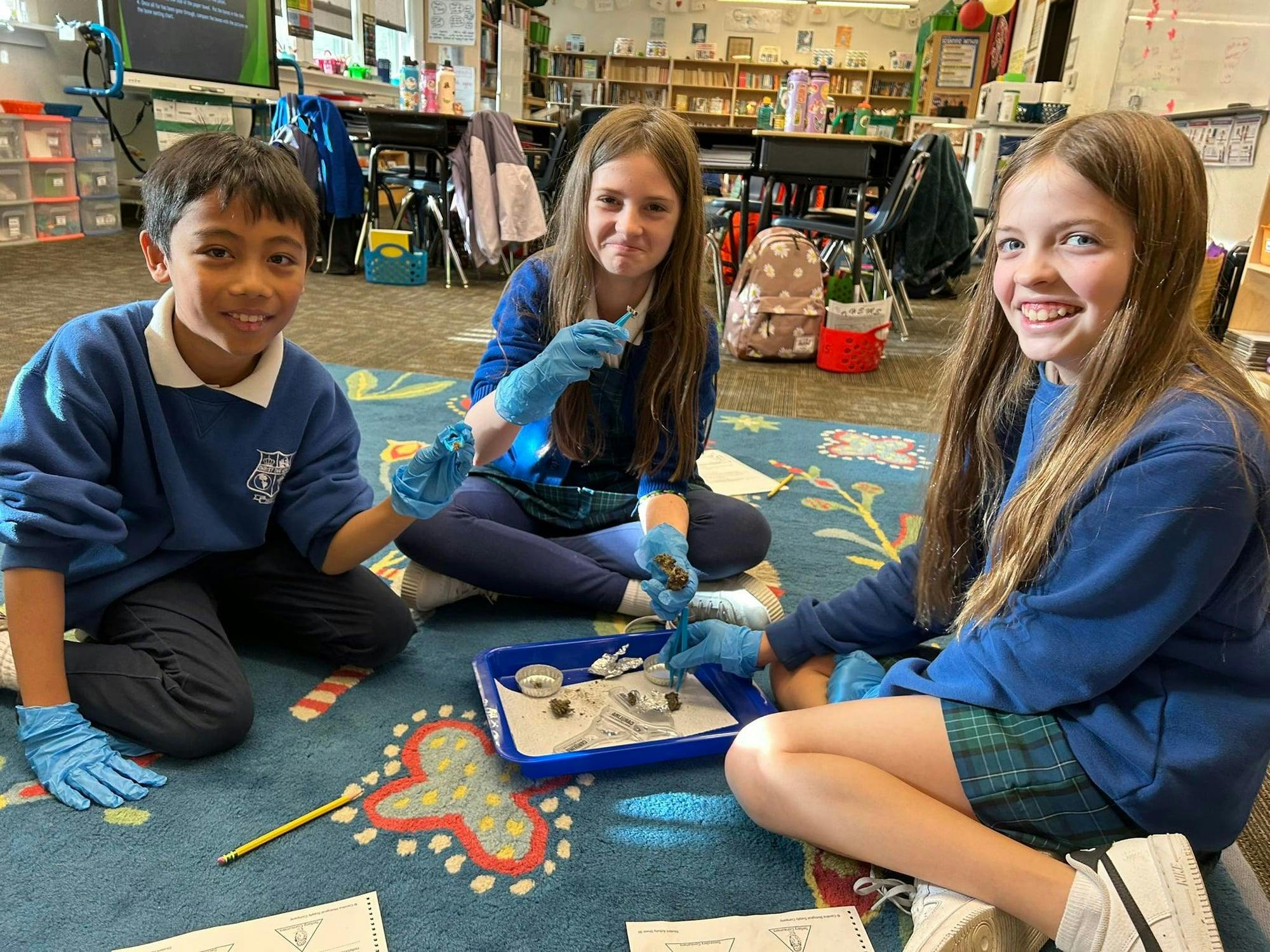 Three children in a classroom examining items on a tray with blue gloves