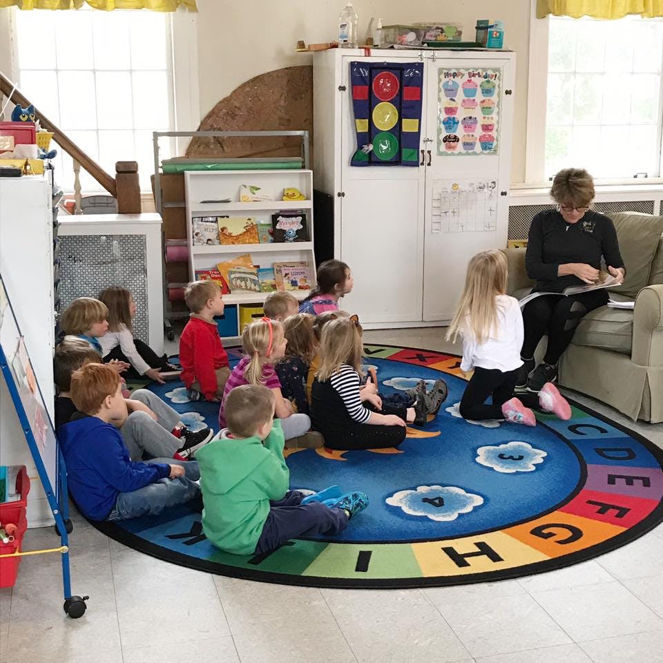 Person reading to a group of children sitting on a rug in a classroom