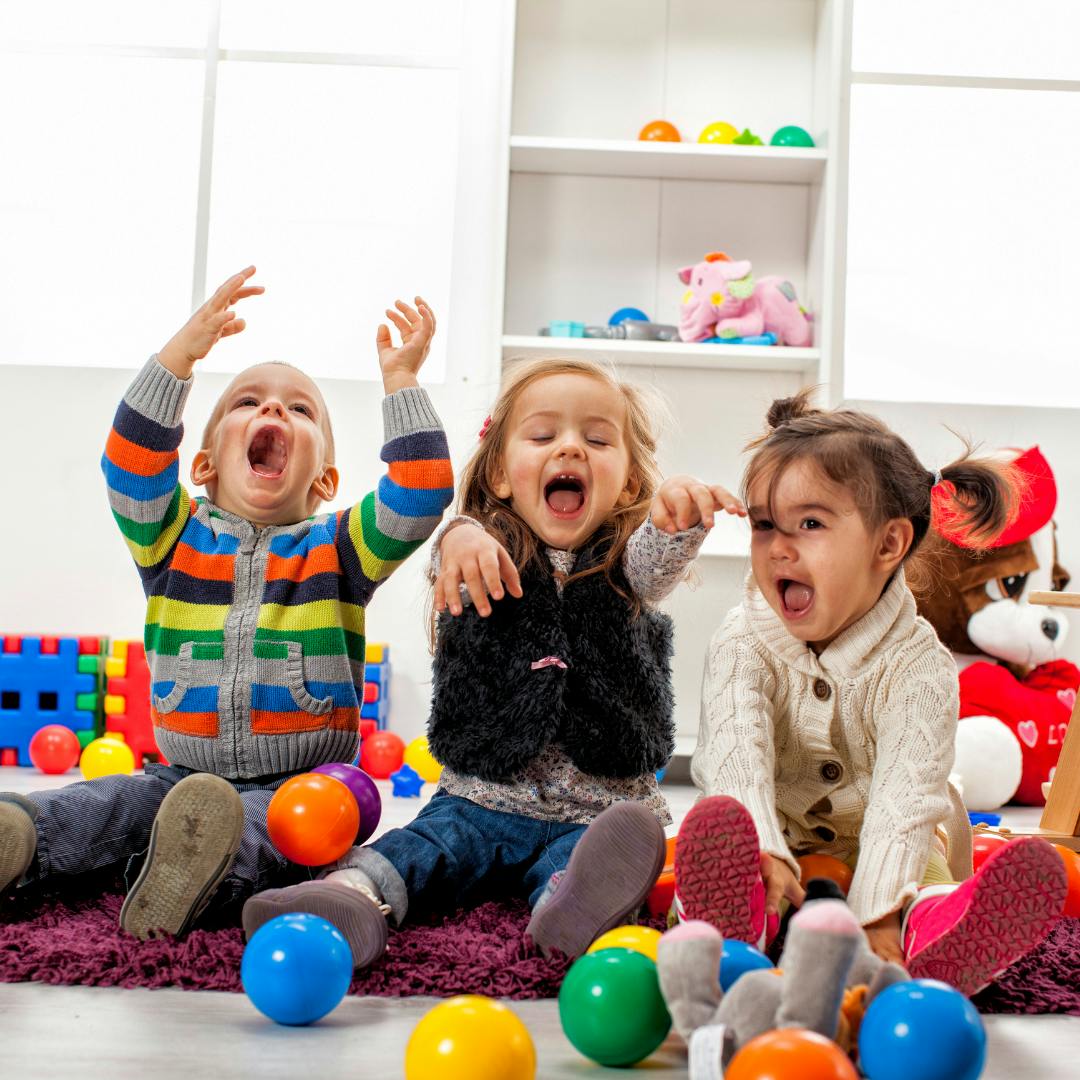 Three children sitting on a rug with open mouths and raised arms