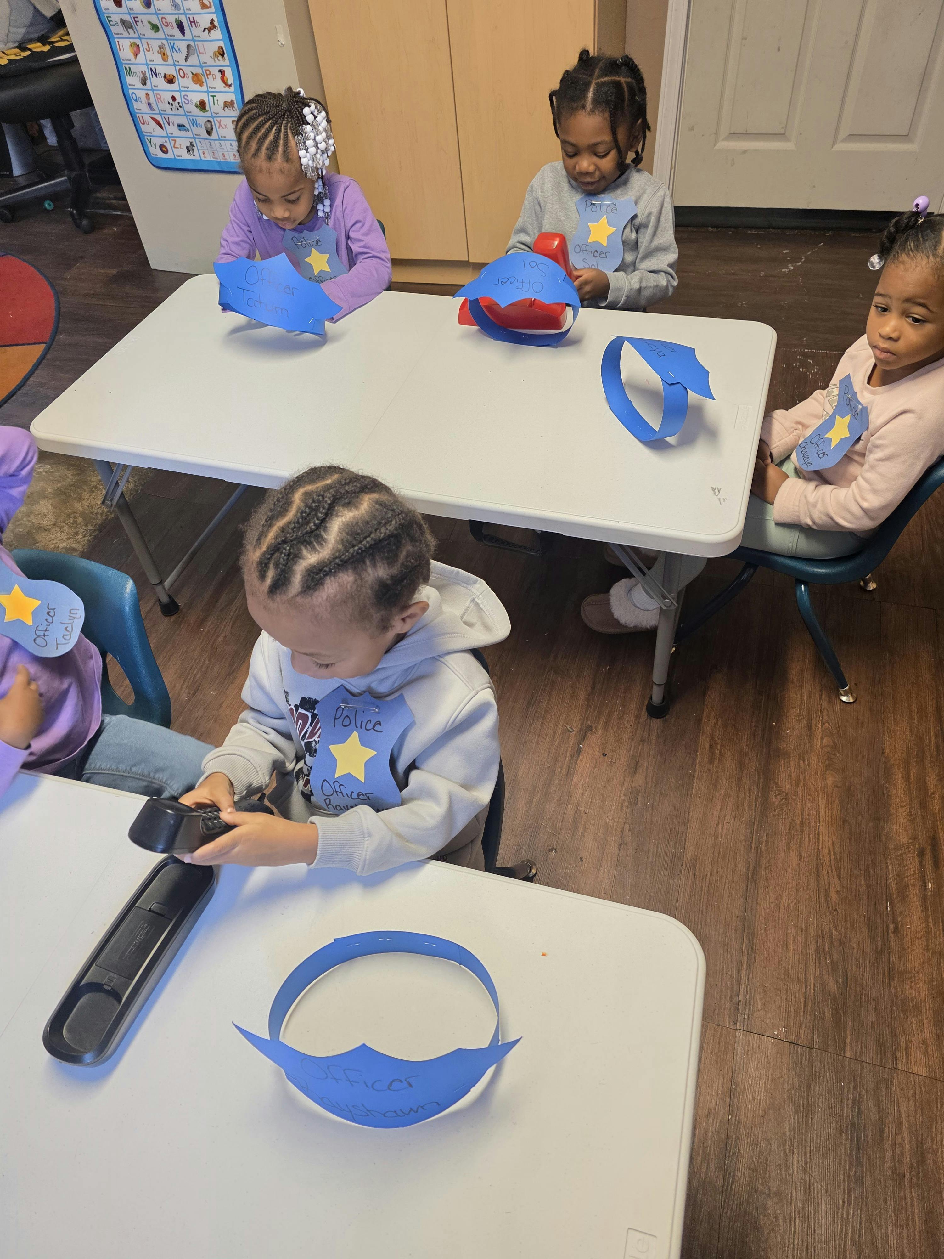 Several children at a table with paper police badges and hats, crowns, and a child holding a toy phone