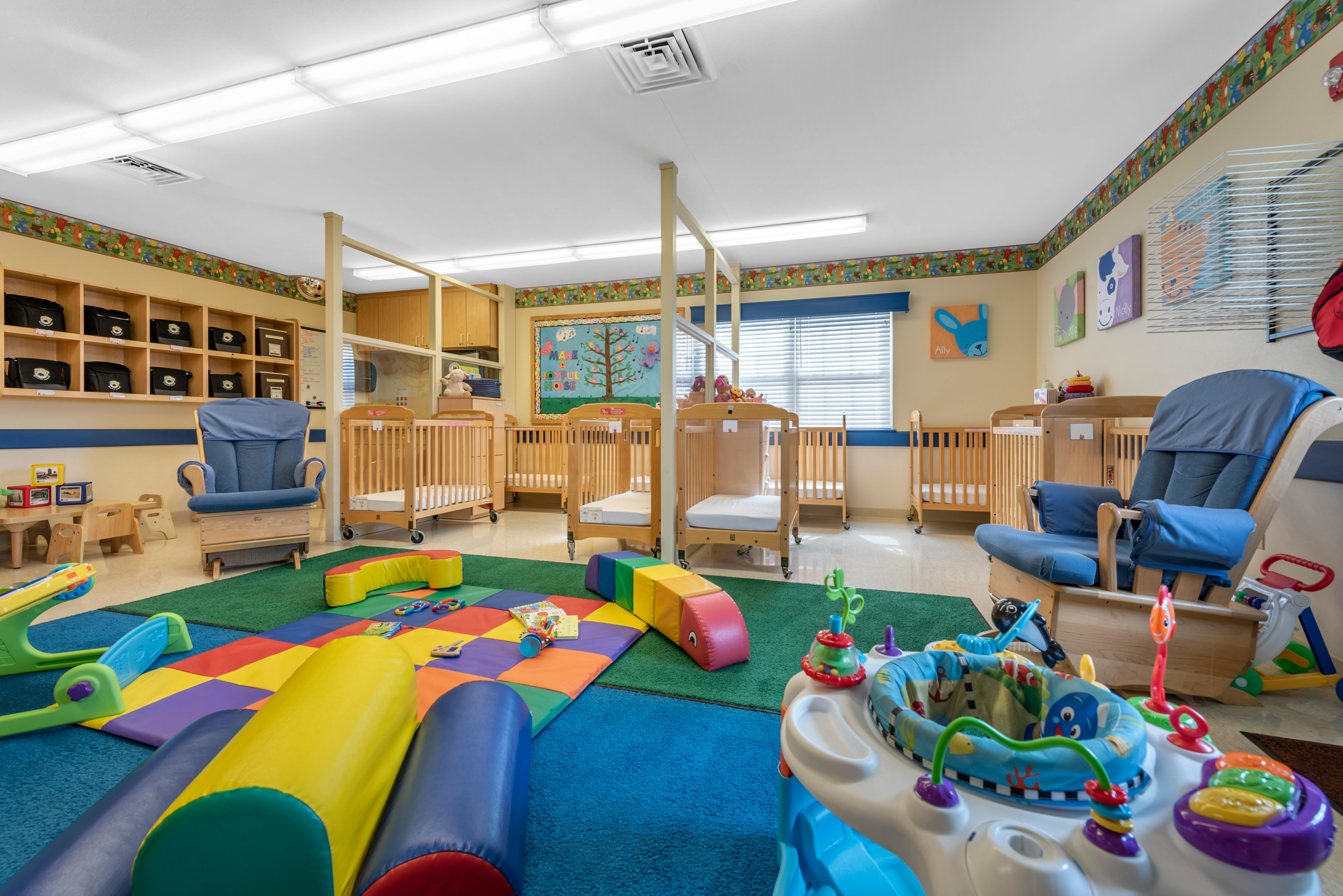 A childcare room with multiple cribs, a blue rocking chair, and colorful play mats on the floor