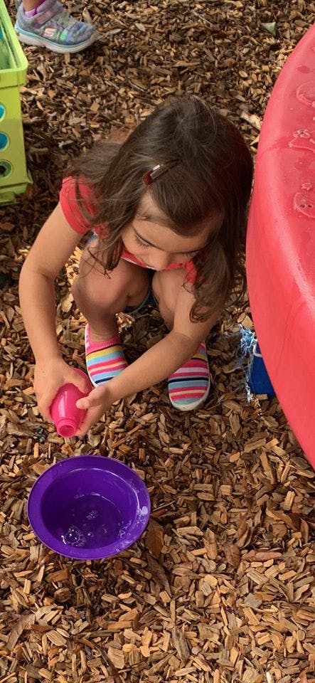 Child crouching and pouring water from a pink cup into a purple bowl