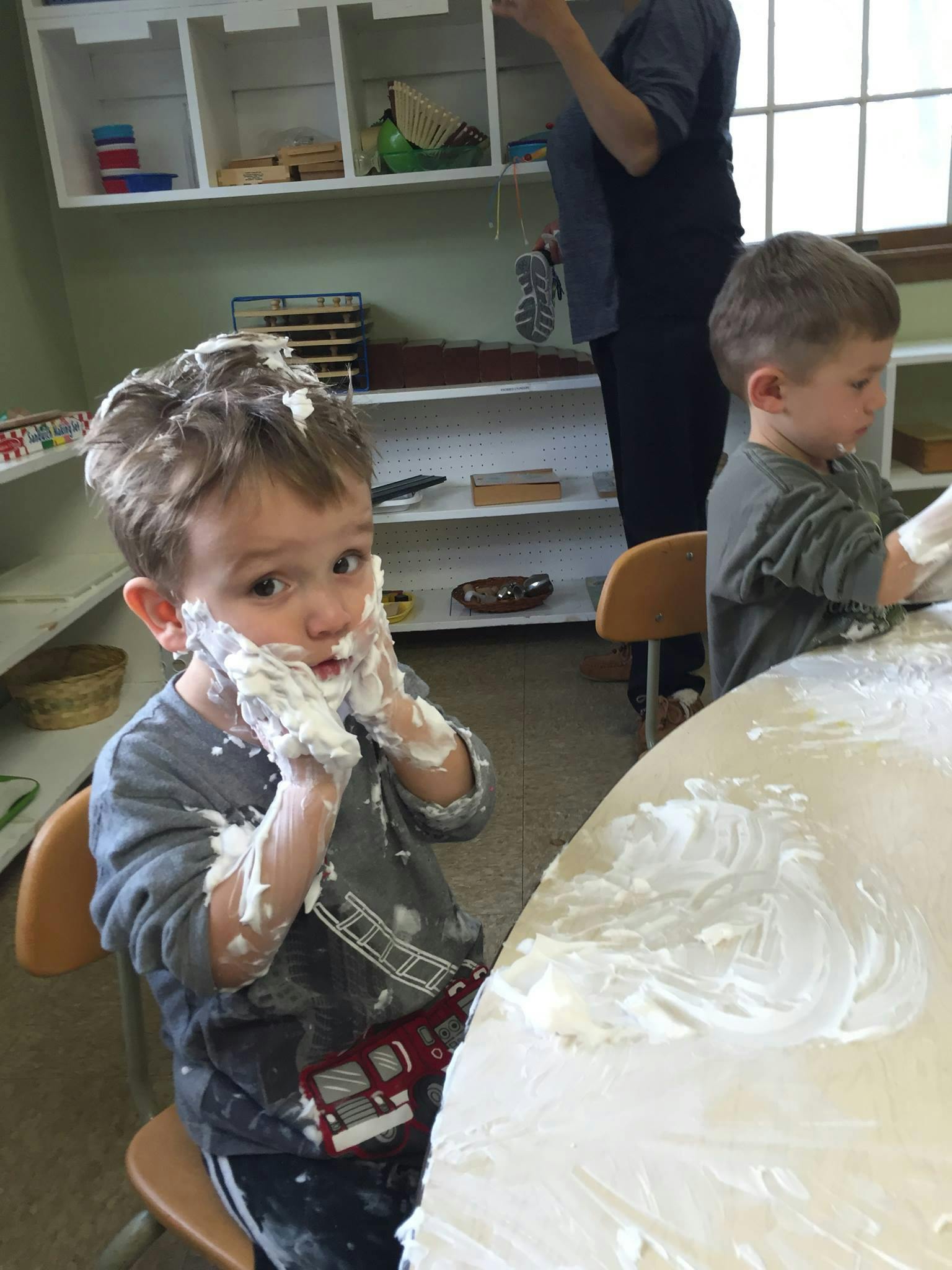 Child with shaving cream on face and hands, another child at a table