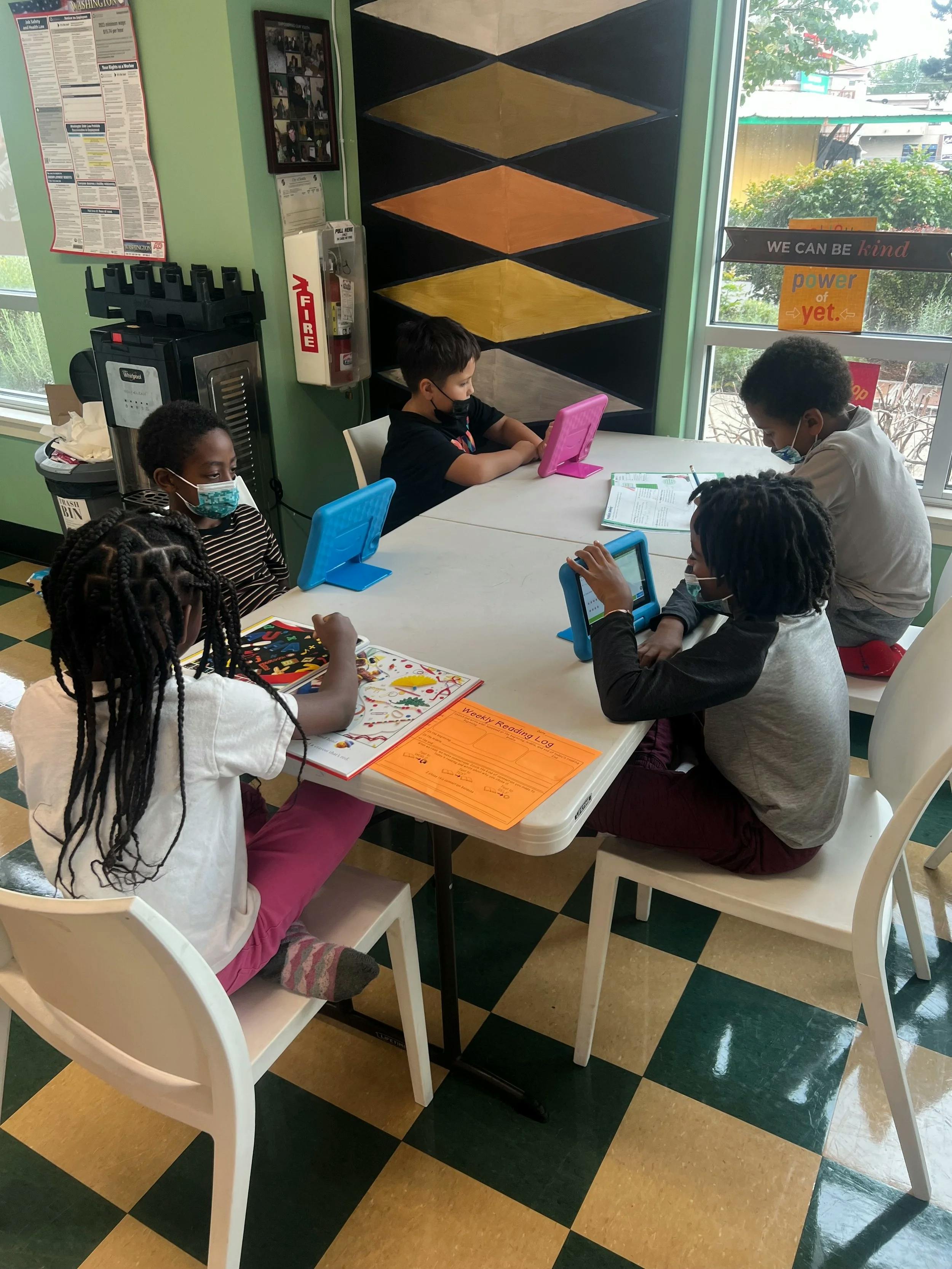 Five children sitting around a table, two reading books and three looking at tablets