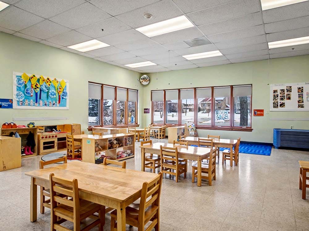 Empty classroom with small wooden tables and chairs, large windows, and art on walls