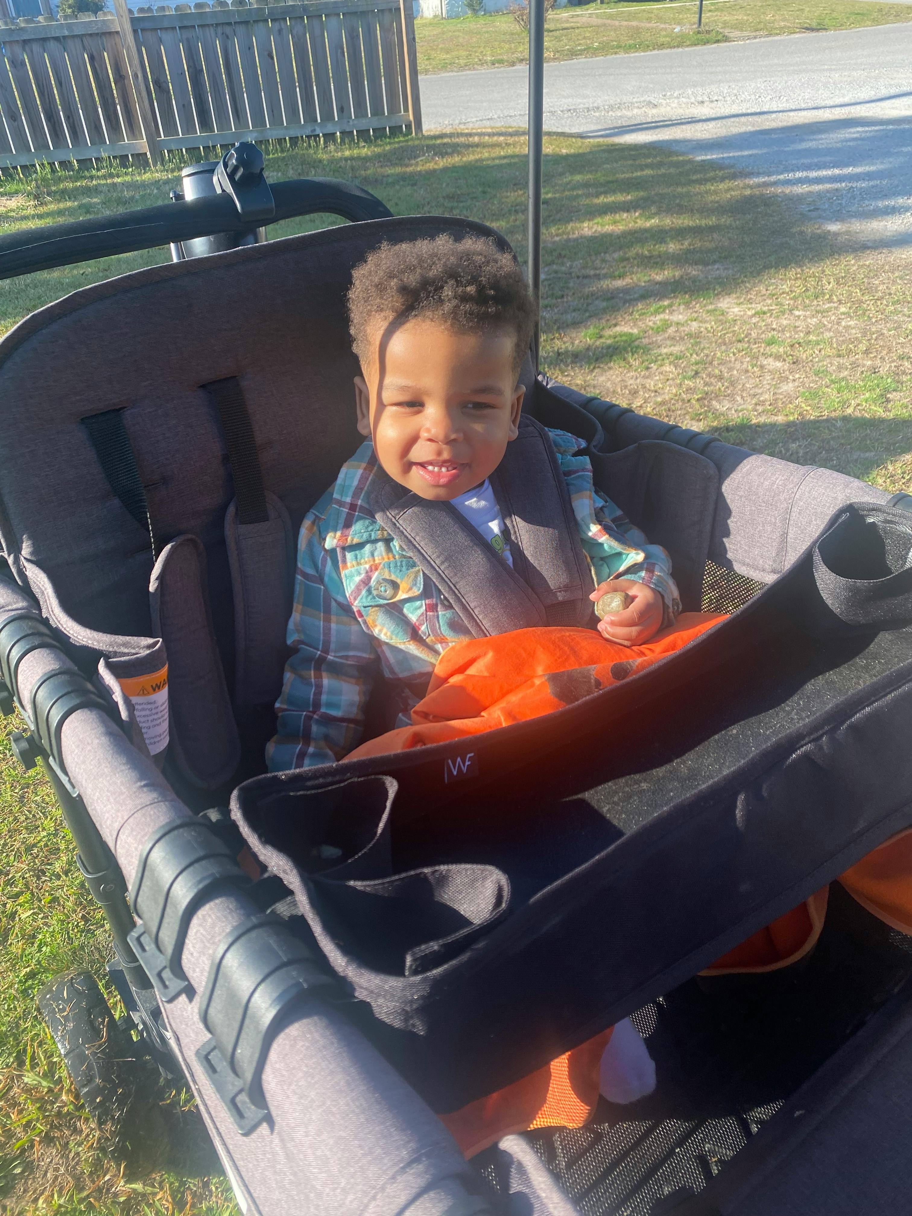 Child smiling in a stroller, wearing a plaid shirt and orange blanket