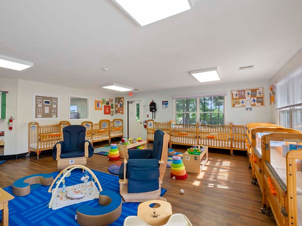A brightly lit nursery room with multiple wooden cribs, chairs, and infant play items