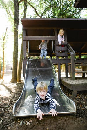 Child sliding down a playground slide, two other children on the play structure