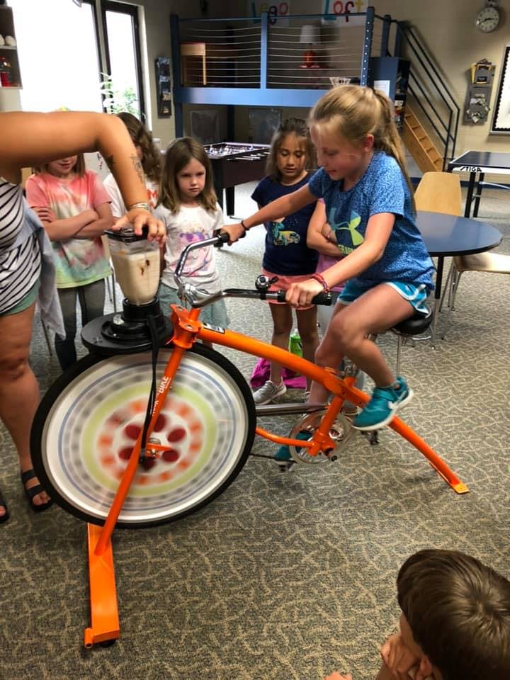 Child on a stationary bike powering a blender with other children watching