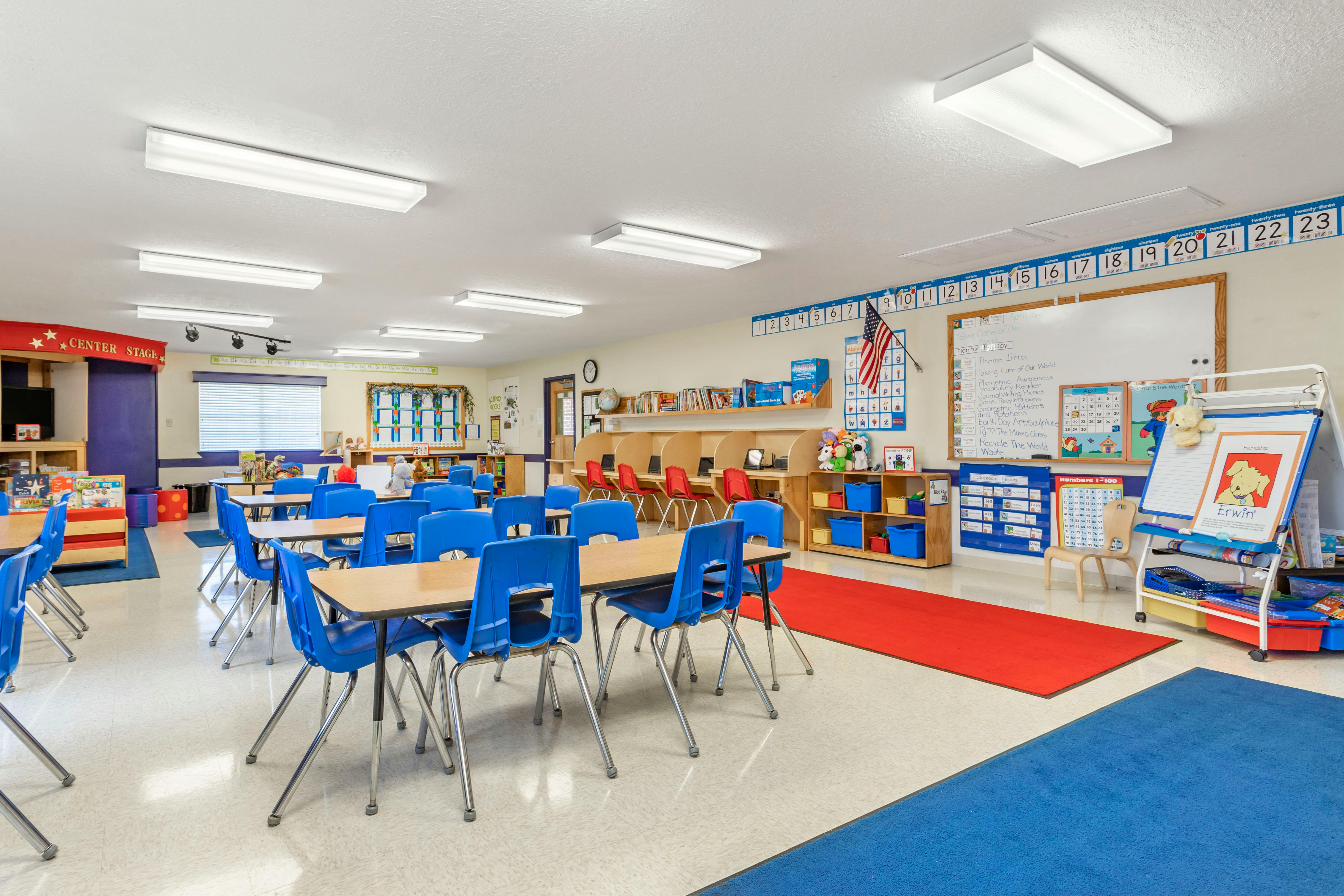 Classroom with small tables and blue chairs, red and blue rugs, and educational posters