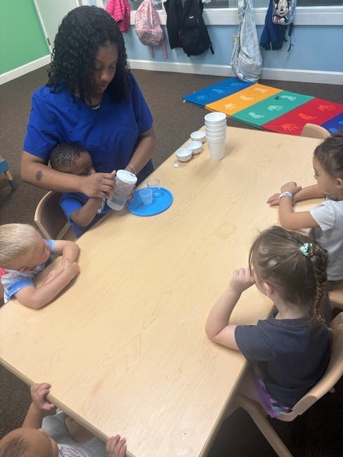 Person helping a child drink from a cup at a table with other children