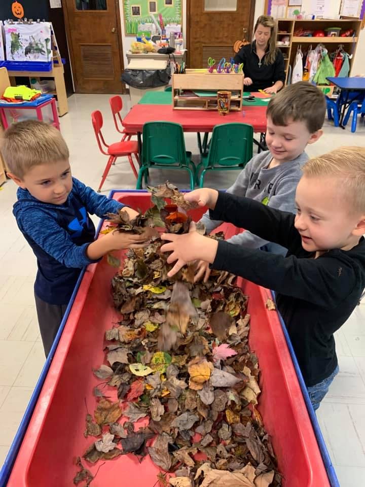 Three children playing with leaves in a red bin in a classroom